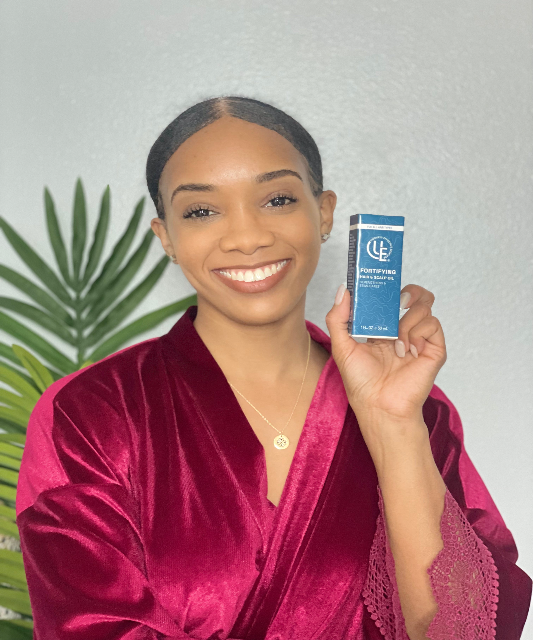 Model holding box with bottle of Fortifying Hair & Scalp Oil from Uniquely Effective Skincare, shown with botanical ingredients to highlight its natural, nourishing formula.