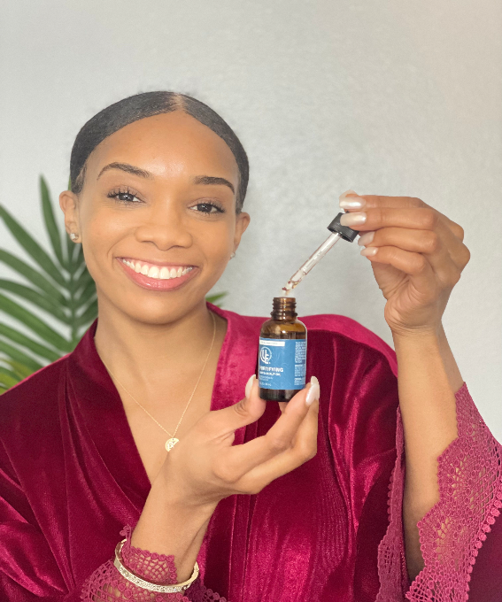 Beautiful model smiling while holding a  glass bottle with dropper of Fortifying Hair & Scalp Oil from Uniquely Effective Skincare, shown with botanical ingredients to highlight its natural, nourishing formula.